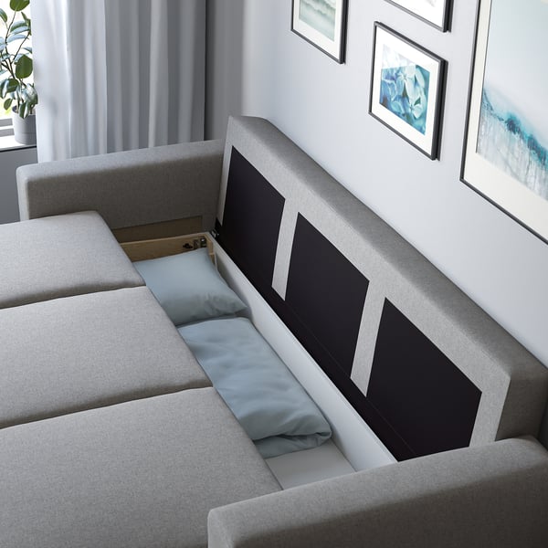 Modern grey sofa-bed with large cushions, featuring a practical storage compartment open, displaying pillows inside.