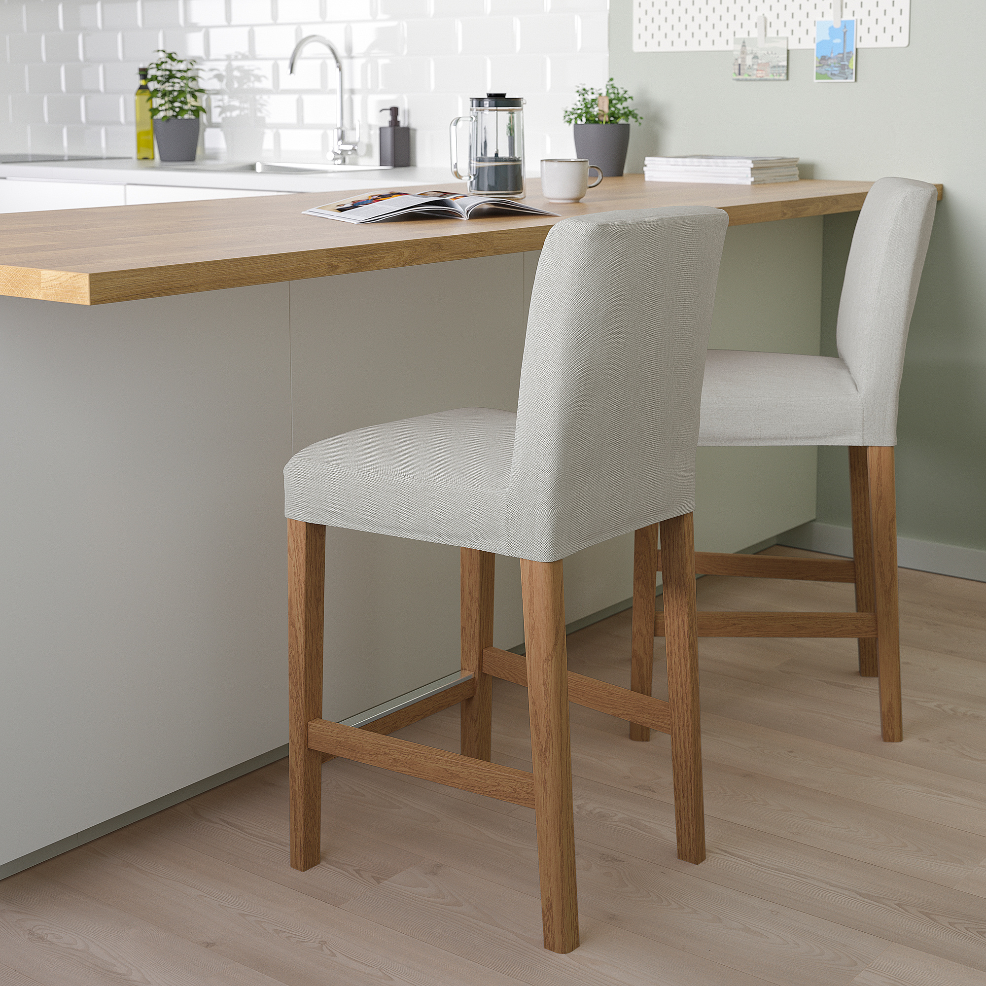 Two grey fabric BERGMUND barstools with wooden legs at a kitchen island, smaller than the potted plants and counter items.