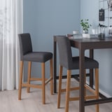 Modern bar stools, named BERGMUND, featuring grey, cushioned seats with wooden legs, positioned beside a high table; sleek and compact design.