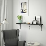 A cozy living area with a BERGSHULT black shelf holding a plant, decor, and a frame. Below, a table holds a book and a glass.
