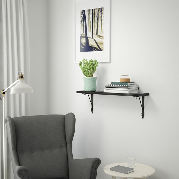 Minimalist room with black BERGSHULT wall shelf. Displaying books, plant, and decor above grey chair. White framed artwork above.