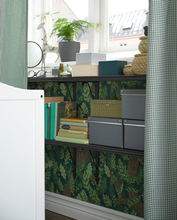 Bookshelf with green leaf-patterned wallpaper, holding books, boxes, and a plant.