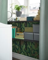 Bookshelf with green leaf-patterned wallpaper, holding books, boxes, and a plant.