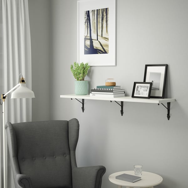 A cozy corner with a gray armchair beside a white wall shelf displaying a plant, books, frames, and a lamp.