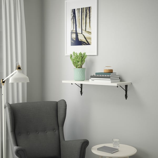 Gray armchair next to small white table. Wall shelf with green plant and books. Art print above.