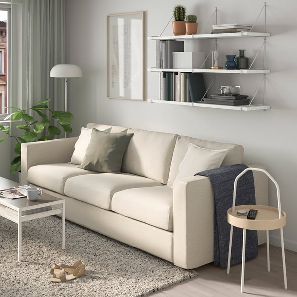 Modern living room with cream BERGSHULT sofa, wall shelf with books and plants, side table with remote and lamp.