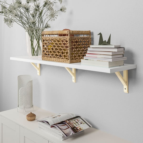 White wall shelf mounted with 3 brackets. Displaying books, woven basket, vase with flowers, and a stack of magazines.