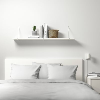 Minimalist bedroom with BERGSHULT shelf above bed, holding books, plant, and basket. White linens on bed, clean modern decor.