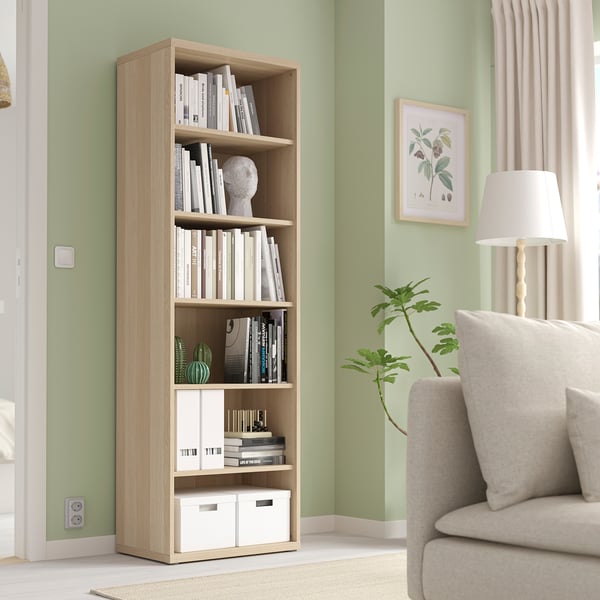 Wooden BESTÅ bookcase with books & decor, taller than chair & lamp.