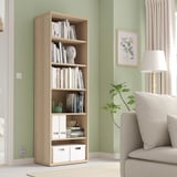Wooden BESTÅ bookcase with books & decor, taller than chair & lamp.