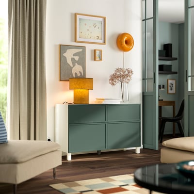 Modern room with teal & white BESTÅ cabinet, lamp, artworks, and neutral curtains, adjacent to a sleek glass door.