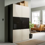 Modern BESTÅ storage unit with glass doors, cream and black, in living space with couch and plant.