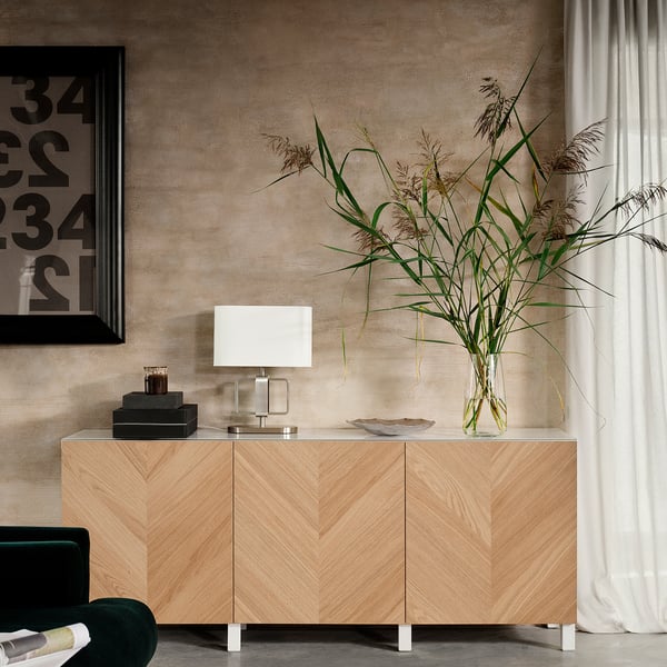 Wooden sideboard, minimalist design, beige finish. White lamp, glass vase with plants, and black framed picture.