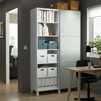 White BESTÅ storage unit with adjustable shelves, storing boxes and books, taller than nearby furniture.