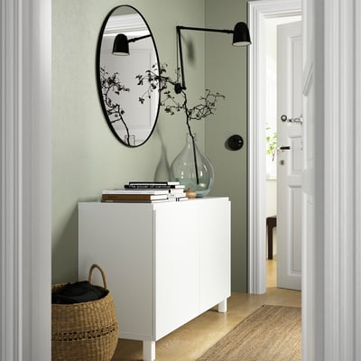 Modern hallway with BESTÅ white sideboard, wide & low, holding books, vase with branches, and mirror above. Basket below, doors open to another room.