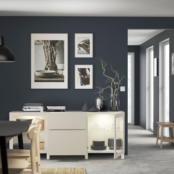 Modern living room with BESTÅ sideboard and lit cabinets showing glassware and plates, plus vases on top, against a gray wall with art prints.