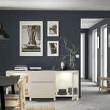Modern living room with BESTÅ sideboard and lit cabinets showing glassware and plates, plus vases on top, against a gray wall with art prints.