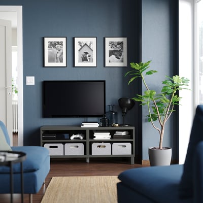 Modern living room with blue accent wall, mounted flat-screen tv on black stand, blue cushioned chairs, house plants, and framed photos.