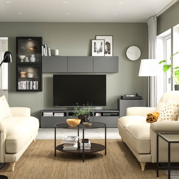 Modern living room with sage green walls, featuring white sofas, a glass coffee table, and a gray BESTÅ tv bench with storage and adjustable shelves.