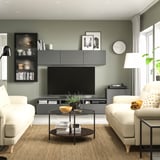 Modern living room with sage green walls, featuring white sofas, a glass coffee table, and a gray BESTÅ tv bench with storage and adjustable shelves.