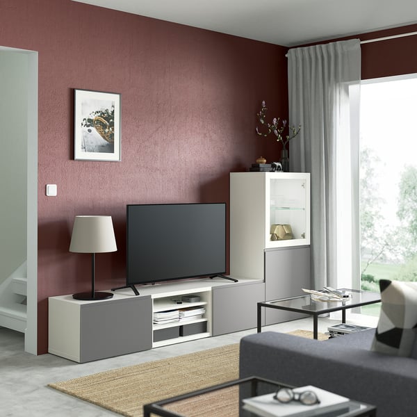 Modern living room with BESTÅ tv stand, flat screen tv, glass coffee table, grey sofa, red accent wall, large window with curtains, and a rug.
