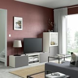 Modern living room with BESTÅ tv stand, flat screen tv, glass coffee table, grey sofa, red accent wall, large window with curtains, and a rug.