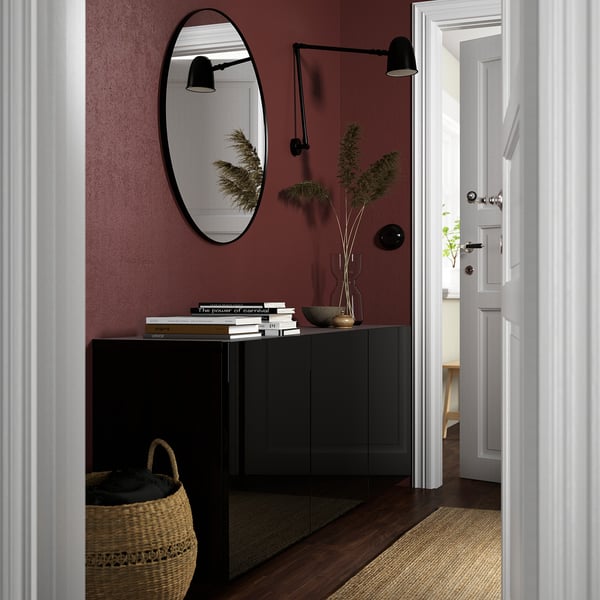 Modern entryway with BESTÅ cabinet, mirror, books, plant. Burgundy wall, wicker basket, and rug create cozy, stylish space.