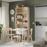 Modern kitchen nook with BILLY bookcase desk, wooden chairs, white table. Green walls, shelves with dishes, glasses, and decor.