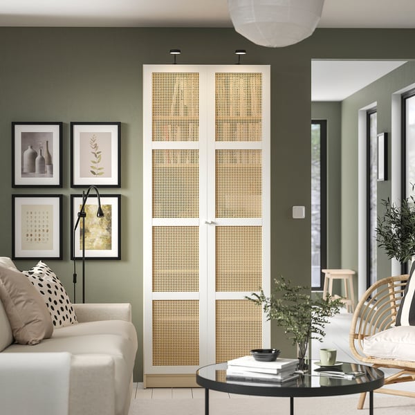 Modern living room with olive walls, white couch, wicker chair, glass table, bookcase, art, hanging lights.