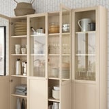 Wooden BILLY bookcase with glass doors, storing dishes, glasses, and is anchored to the wall.