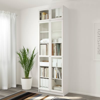 A tall white BILLY bookcase with glass doors, holding books and decor, against a white wall. A large plant sits beside it.