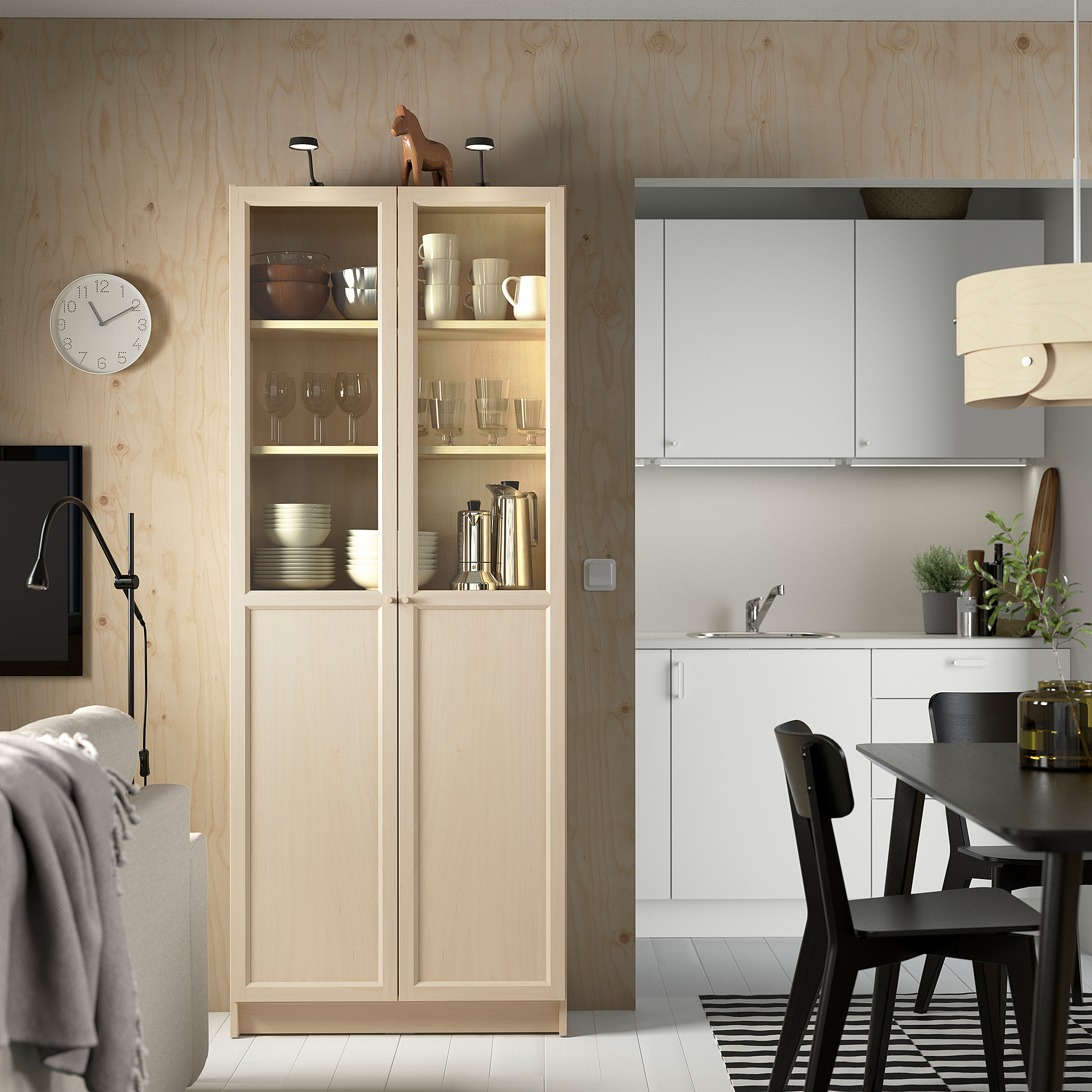 Modern kitchen with BILLY light-yellow cabinet displaying dishes, beside black dining set. White kitchen area has sink and plants.