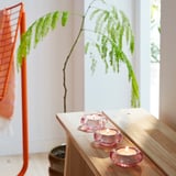 Image with three pink glass BLADFÅGEL tealight holders on a wooden shelf, each lit, near a large green fern and an orange chair.