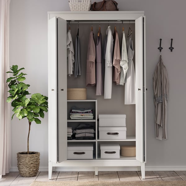 Open white wardrobe displays clothes and storage boxes, with a brown handbag on top. To the left, a potted plant in a wicker basket.