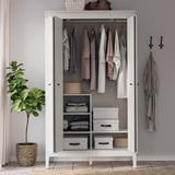 Open white wardrobe displays clothes and storage boxes, with a brown handbag on top. To the left, a potted plant in a wicker basket.