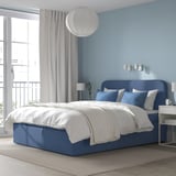 Modern blue ottoman bed with white bedding, large window, grey curtains, and wall lights. Cozy and spacious with a comfortable headboard.