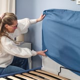 Woman opening BLÅFJÄLLET ottoman bed, revealing storage space.