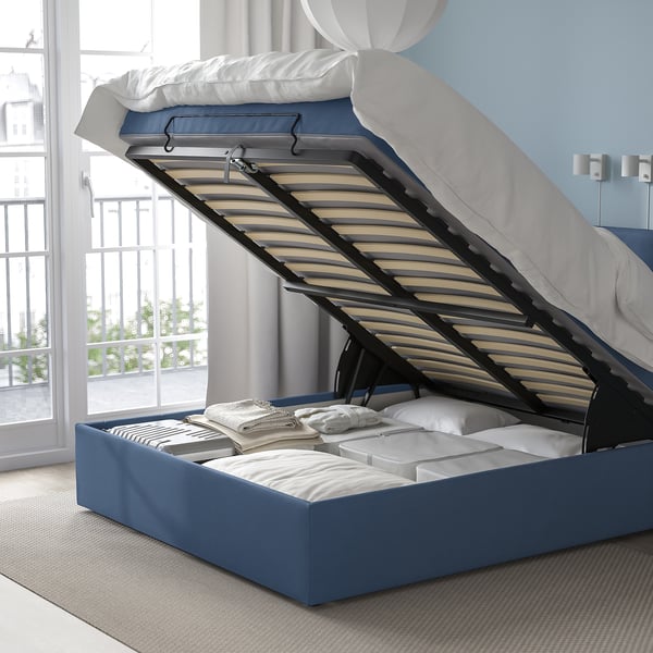 Bed with open storage compartment, revealing slatted base and folded linens, showcasing storage functionality.