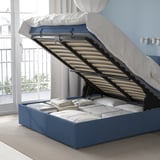 Bed with open storage compartment, revealing slatted base and folded linens, showcasing storage functionality.