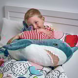 A young boy cuddles a plush blue shark stuffed animal while sitting on a bed. The shark is large and soft, perfect for hugging and play.
