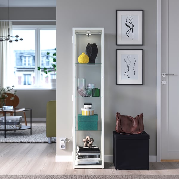 White cabinet w/glass doors, books, spotlight. Brown bag on cube, prints, green couch.