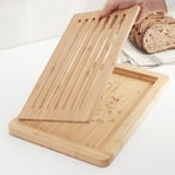 Image shows a bamboo chopping board with grooves to collect crumbs. A hand lifts the tray to empty crumbs, highlighting its functionality.