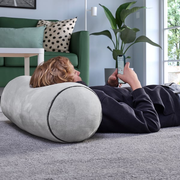 A person lies on a cylindrical cushion, using a smartphone. The cozy setting includes a green couch and a plant.