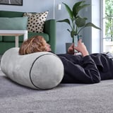A person lies on a cylindrical cushion, using a smartphone. The cozy setting includes a green couch and a plant.