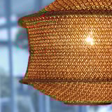 Close-up of intricate woven pendant lamp, casting warm light. Golden-hued with detailed mesh pattern.