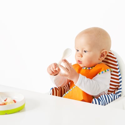 Baby in high chair eats with spoon. Spoon has grip-friendly design, suitable for learning self-feeding.