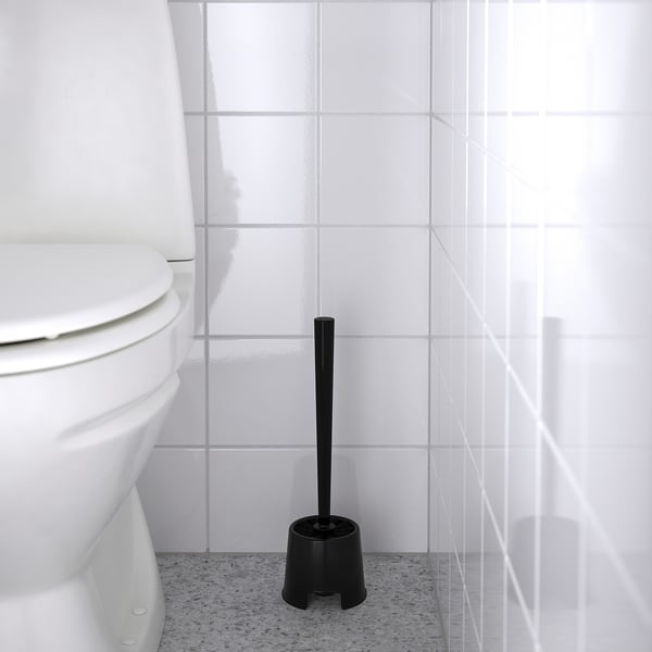 Black toilet brush in holder beside white toilet. Cleaning tool stored upright on speckled grey floor.