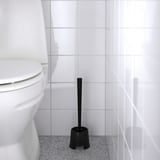 Black toilet brush in holder beside white toilet. Cleaning tool stored upright on speckled grey floor.