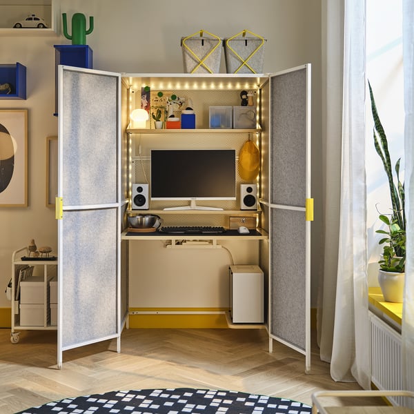 Gaming station with monitor, speakers, keyboard, and decorations on shelves, lit with fairy lights, placed in a room with a plant and a checkered rug.