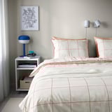 Modern bedroom with BREDVECKLARE bedding in greige and red, white bedspread, matching pillows, side table, and geometric wall print.
